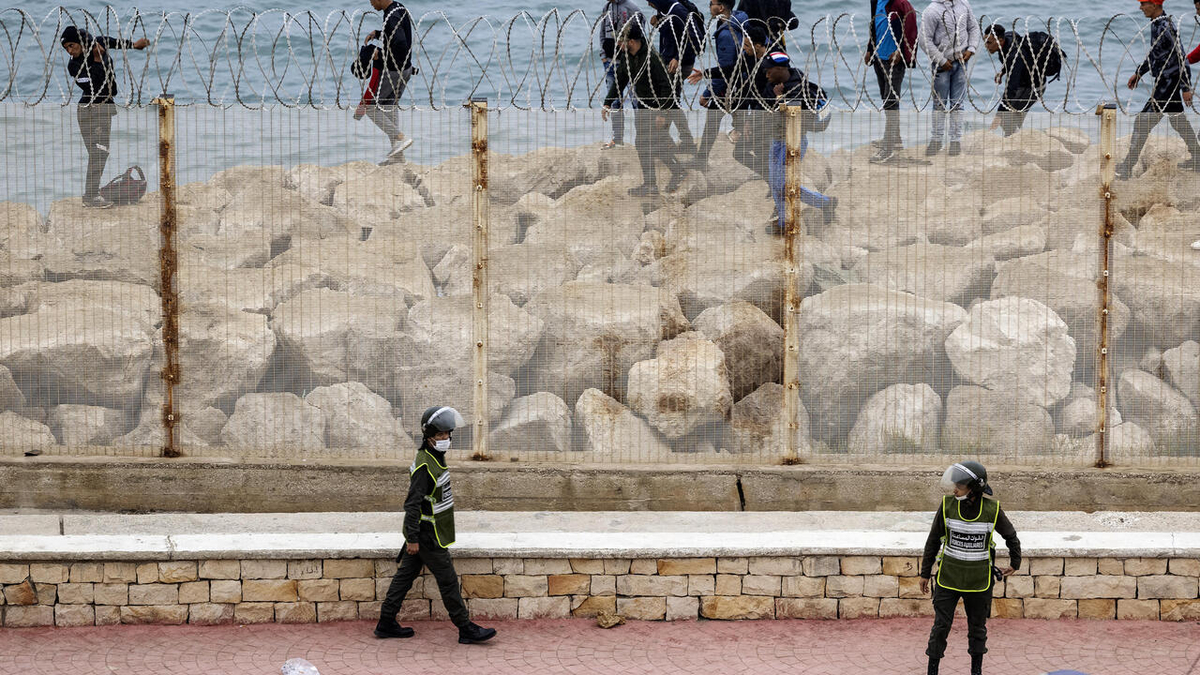 Moroccan security officers stand guard as migrants walk by the shoreline in the northern town of Fnideq in an attempt to cross the border from Morocco to Spain's North African enclave of Ceuta on May 18, 2021. - At least 5,000 migrants, an unprecedented influx at a time of high tension between Madrid and Rabat, slipped into Ceuta on May 17, a record for a single day, Spanish authorities said. They reached the enclave by swimming or by walking at low tide from beaches a few kilometres to the south, some using inflatable swimming rings and rubber dinghies. (Photo by FADEL SENNA / AFP)